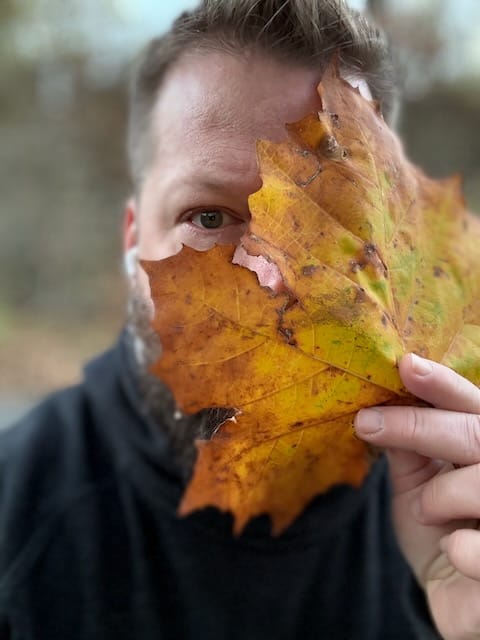 John Paul Davis holding a very large yellow and orange maple leaf in front of half his face